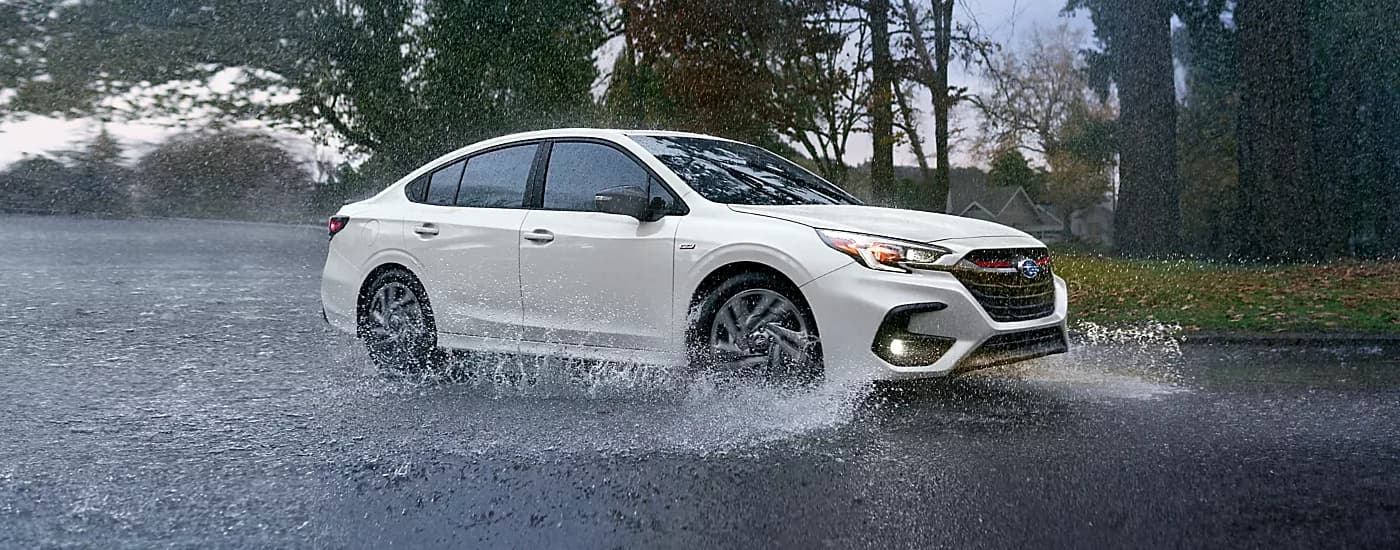 A white 2023 Subaru Legacy splashing through a puddle on a rainy street.