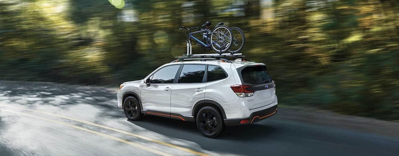 A white 2021 Certified Pre-Owned Subaru Forester Sport is shown with bikes on the roof driving down a blurred road.