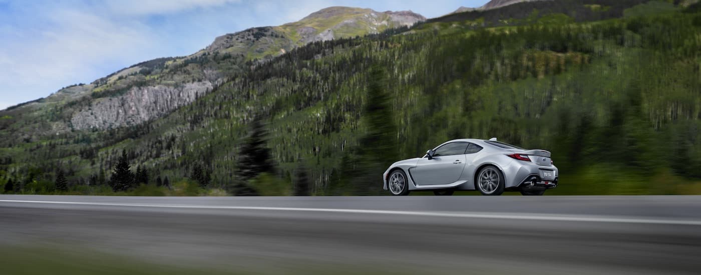 A silver 2022 Subaru BRZ is shown driving past trees and mountains.