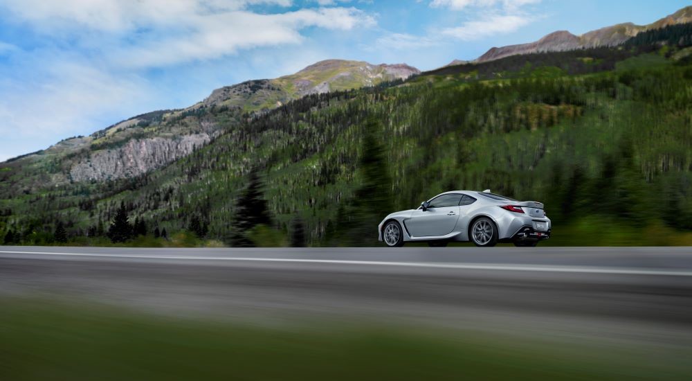 A silver 2023 Subaru BRZ driving on an open road past green mountains.