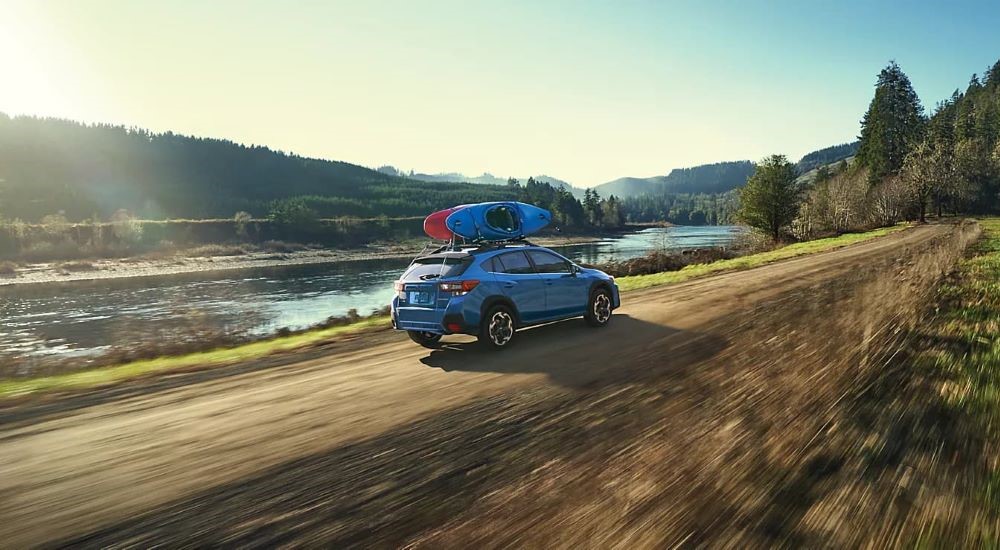 A blue 2023 Subaru Crosstrek with kayaks on the roof driving near a river.