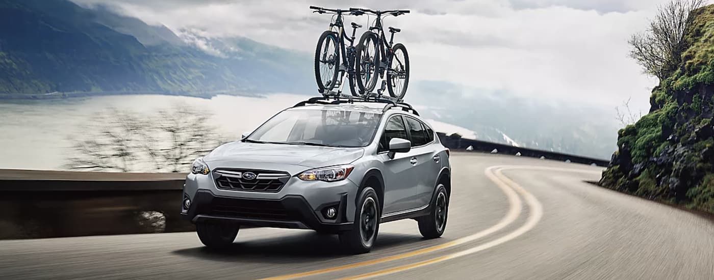 A silver 2023 Subaru Crosstrek Premium with bikes on the roof driving past a lake.