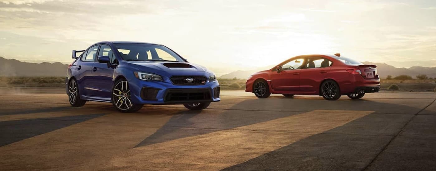 A blue and a red 2021 Subaru WRX STI are parked in front of desert mountains at sunset.
