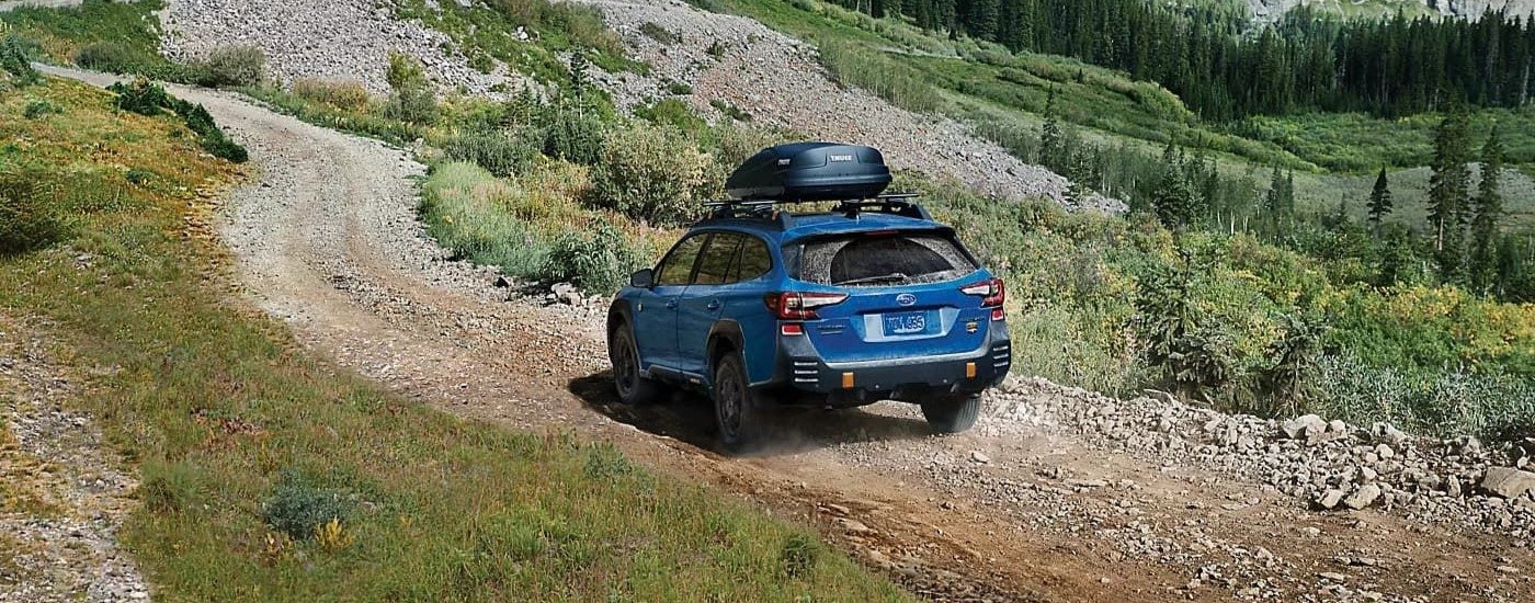 A blue 2023 Subaru Outback Wilderness is shown from the rear on a tree-lined trail.