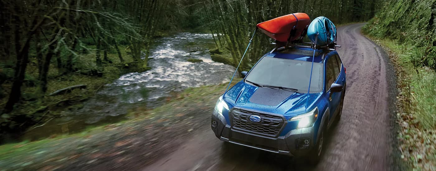 A blue 2023 Subaru Forester Wilderness is shown with kayaks on the roof driving on a dirt road.