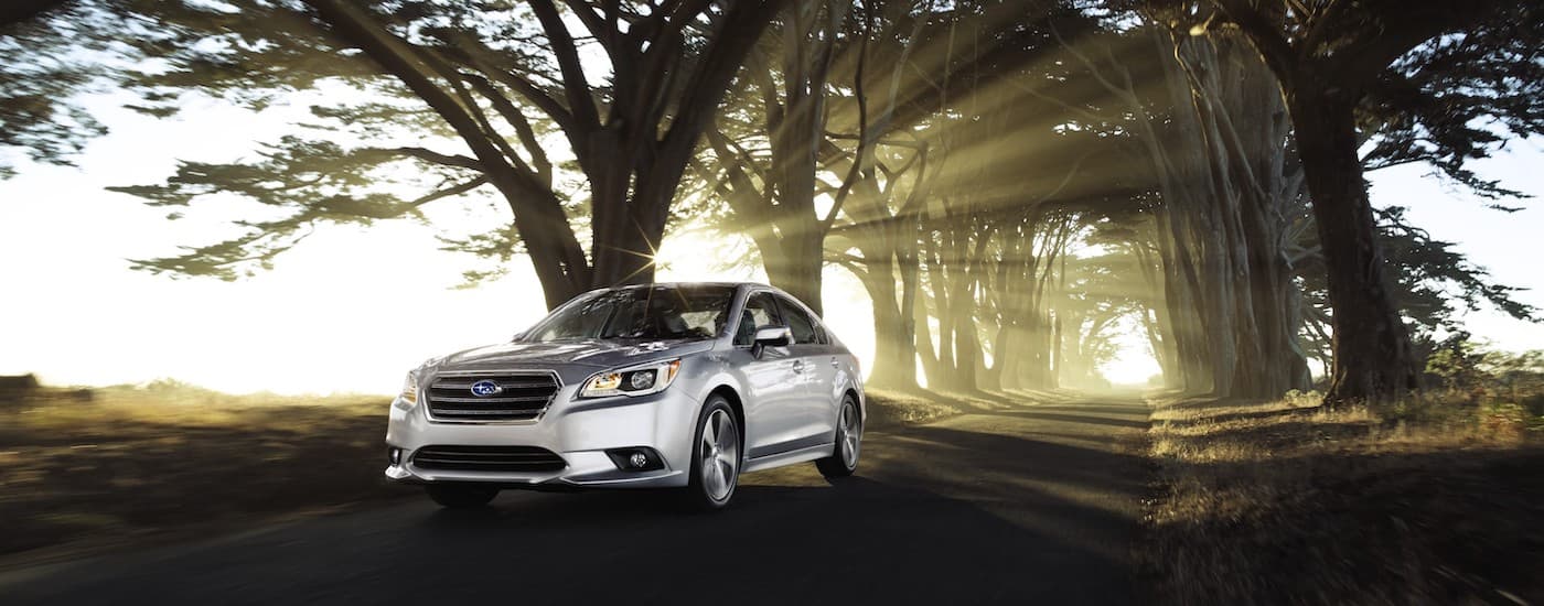 A silver 2017 Subaru Legacy is shown driving down a wooded road with sunlight shining through the trees.